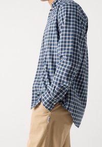 Mężczyzna w niebiesko-zielonej koszuli w kratę z długim rękawem, luźno włożonej do beżowych spodni, ze rękami w kieszeniach, zwrócony bokiem.