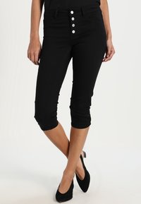 Musta capri-farkut, joissa on korkea vyötärö, viiden napin vetoketju ja kapea siluetti, jotka sisältävät käänteet haalareissa ja sileän denim-tekstuuri.