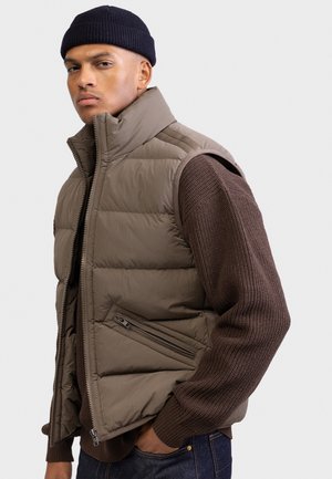 Ung mand iført en mørk hue, brun strikket sweater og en brun polstret vest med en lynlåslomme, står med den ene hånd i lommen.