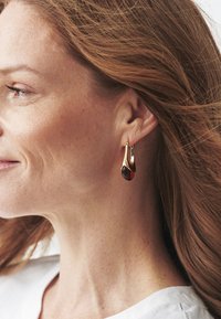 Náušnice v zlatej farbe s plynulým kvapkovým dizajnom a hlbokým červeným akcentovým kameňom, ktoré majú hladké krivky a lesklý povrch.