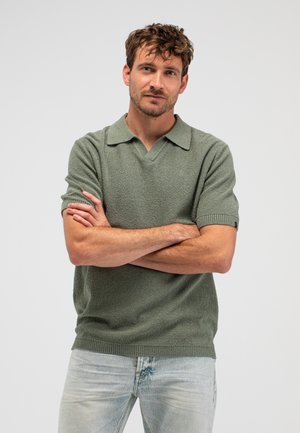 Mann mit kurzen lockigen Haaren, der einen grünen Kurzarm-Pullover und hellblaue Jeans trägt, steht mit verschränkten Armen vor einem einfarbigen Hintergrund.