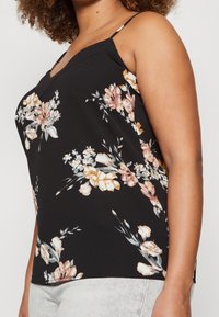 Svart blommig camisole med V-ringning, tunna axelremmar och en blandning av rosa, beige och vita blommor med gröna blad. Slät tygtextur.