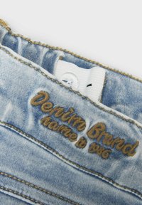 Svijetloplava traper tkanina s izblijeđenom teksturom. Sadrži smeđi vezeni natpis "Denim Brand" i bijelu oznaku s gumbom na struku.
