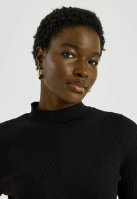 Suéter de punto acanalado negro con cuello alto y líneas verticales, con un patrón sutil. Tono de piel oscuro, con cabello corto y texturizado.