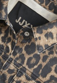 Hedvábná košile s leopardím vzorem zblízka, kombinující odstíny tan, hnědé a černé. Výrazný stříbrný patent a viditelná etiketa uvnitř límce.