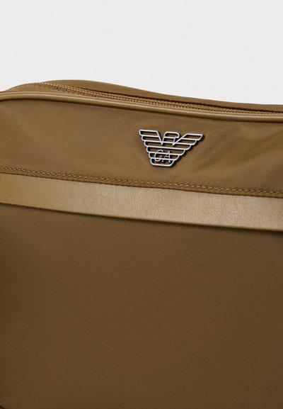 Καφέ υφασμάτινη θήκη με οριζόντια λωρίδα από δέρμα και μεταλλικό λογότυπο αετού με στυλιζαρισμένα φτερά και τα γράμματα GA.