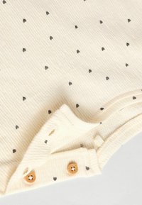 Tela de algodón color crema con un patrón texturizado de ribetes, adornada con pequeños diseños de corazones negros. Presenta botones de madera cerca del dobladillo.
