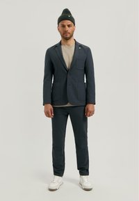 Sacou marin cu buzunare mari, închidere cu butoane, asortat cu pantaloni corespunzători și o cămașă bej deschis dedesubt; modelul poartă o căciulă verde.