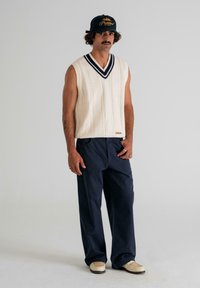 Man wearing cream cable-knit sleeveless sweater, navy wide-leg pants, cream woven loafers, and a black cap with yellow embroidery, standing.