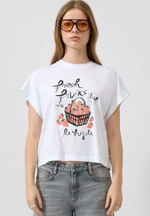Jeune femme portant de grandes lunettes de soleil ambre, un crop top blanc avec un motif panier de fraises et du texte, et un jean gris, se tenant devant un fond uni.