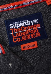 Černá texturovaná látka s námořnickou modrou etiketou, na které je v bílé a oranžové barvě uvedeno "Superdry" a "The Orange Label Polo Co." Velikostní štítek uvádí "STŘEDNÍ".