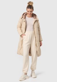 Beige pufferjack met capuchon en rits, met een gequilt ontwerp en tonal voering, gecombineerd met crèmekleurige joggingsbroek en sneakers.