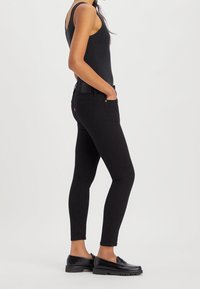 Mujer luciendo una camiseta sin mangas negra, jeans ajustados negros y mocasines negros, de pie en perfil lateral con las manos en los bolsillos.