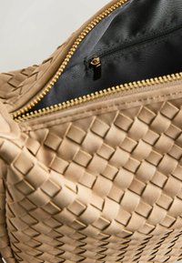 Beige geweven handtas met een textuurpatroon, voorzien van een gouden ritssluiting en een zwarte voering aan de binnenkant, met een klein binnenvak met rits.