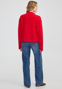 Red pullover with a high neckline, relaxed fit, and long sleeves, paired with blue high-waisted denim jeans. Textured fabric with visible stitching.