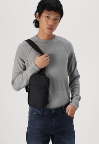 Čierna crossbody taška s obdĺžnikovým tvarom, hladkým povrchom a prednou vreckom na zips. Modelka oblečená v šedom textúrovanom svetri a modrých džínsoch.