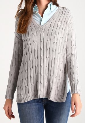 Femme portant une chemise bleu clair à boutons sous un pull gris clair en maille torsadée à col en V, associée à un jean bleu.