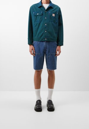 Mężczyzna w zielonej zapinanej na guziki kurtce, przetartych niebieskich dżinsowych szortach, białych skarpetkach i czarnych skórzanych mokasynach na tle jednolitej białej ściany.