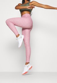 Leggings rosa melangiati con vita alta, abbinati a un reggiseno sportivo verde e scarpe bianche con accenti arancioni.