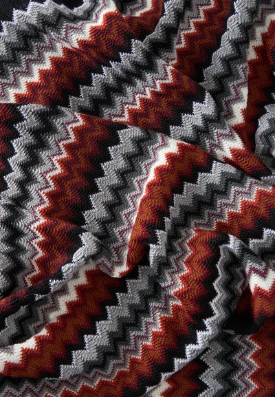 Tissu tricoté doux avec motif en zigzag en rouge, gris, noir et blanc, montrant des plis et la texture en gros plan.