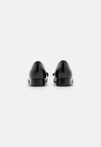 Chaussures en cuir verni noir brillant avec un petit talon carré, ornées d'un accent en nœud à l'avant. Texture lisse et silhouette classique.
