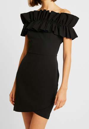 Žena v černých bardot šatech s volánkovým výstřihem a asymetrickým lemem, zobrazená od krku po polovinu stehen.