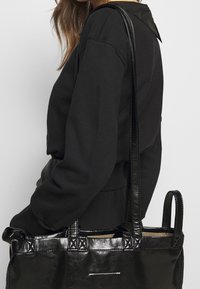 Černá kožená taška přes rameno s dlouhými rukojeťmi, s texturovaným povrchem, strukturovaným tvarem a dekorativními detaily na rukojeti.