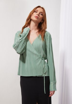 Green, pleated wrap blouse with long sleeves and a tie detail at the waist. Features a deep V-neckline and button accents on sleeves.