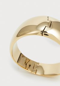 Bracciale in metallo color oro con finitura lucida, dotato di chiusura a cerniera e una sottile incisione di "Calvin Klein". Design curvo e liscio.