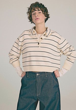 Cream sweater with black stripes, cropped design, buttoned collar, and ribbed cuffs, paired with high-waisted denim jeans.