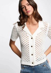 Cardigan bianco a maniche corte con motivo a maglia aperta, design con colletto e dettagli neri sui bottoni, abbinato a pantaloni in denim blue.