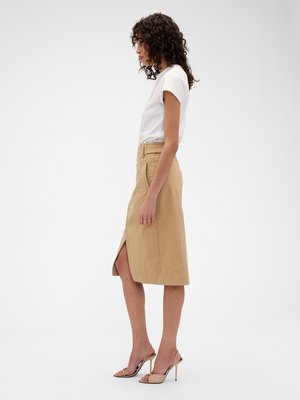 Frau steht seitlich, trägt ein weißes kurzärmeliges Oberteil, einen beigen knielangen Rock mit seitlichem Schlitz und hautfarbene Sandalen mit hohen Absätzen.