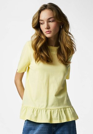 Młoda kobieta o falowanych blond włosach, ubrana w bladoróżowy bluzkę z krótkim rękawem i falbanką na dole oraz niebieskie dżinsy, patrząca w dół na bok.