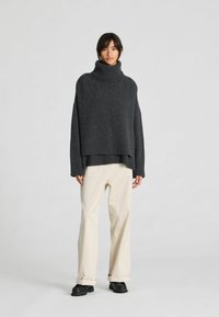 Naine kannab tumedat halli lõdva kõrge kaelusega lõngasärki, kreemikaid laia jalaga pükse mansettidega ning musti kingi, seistes ühtlase tausta ees.