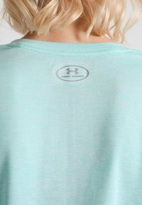 Blond person som bär en ljusblå skjorta med en Under Armour-logotyp tryckt på övre ryggen, synlig under nacken.