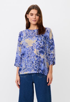 Femme debout face à l'avant, portant un chemisier ample beige à motif floral bleu et un jean bleu large, sur un fond clair uni.