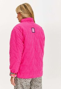 Różowa kurtka z kordurojowego materiału z wysokim kołnierzem, o pikowanej strukturze i czarną metką z tyłu, w połączeniu z wzorzystymi rękawami oraz spodniami w panterkę.