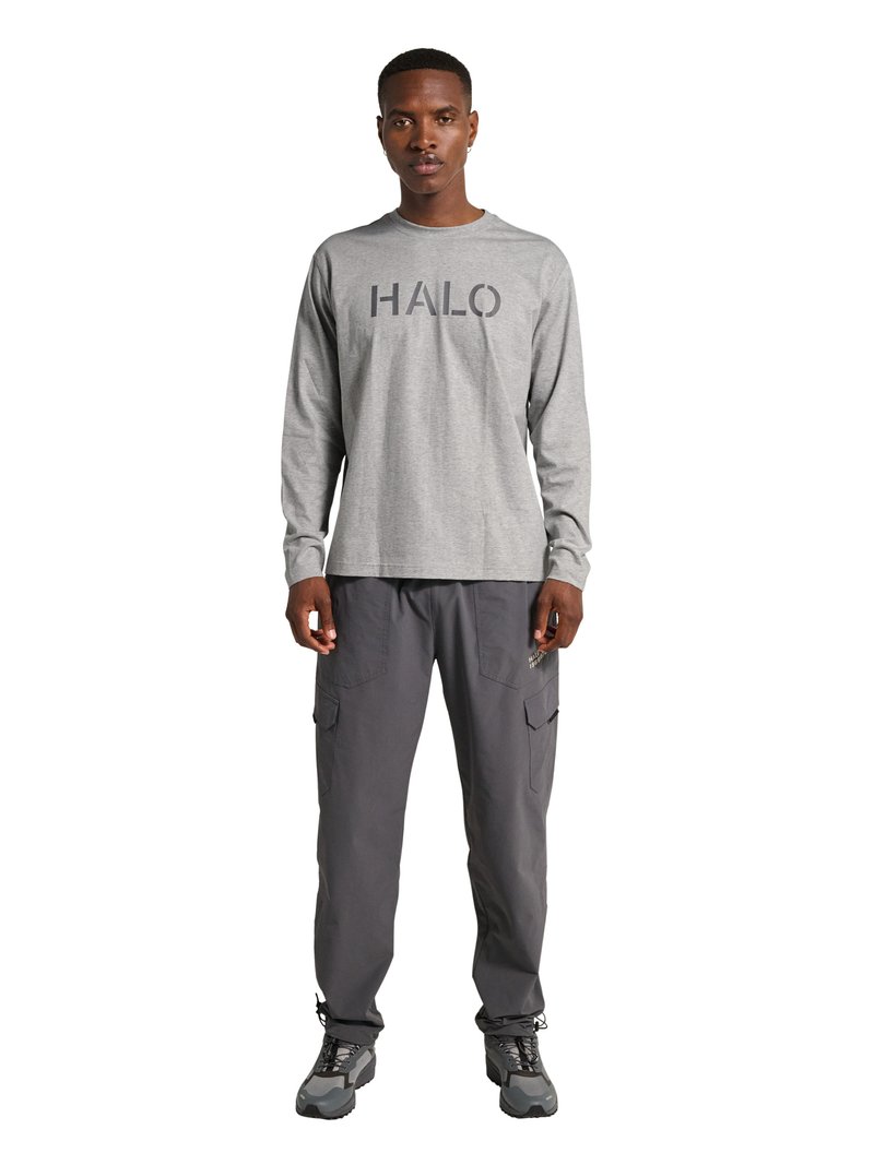 Man draagt een grijs shirt met lange mouwen met de tekst "HALO", donkergrijze cargobroek en zwart-grijze wandelschoenen, staand met het gezicht naar voren.