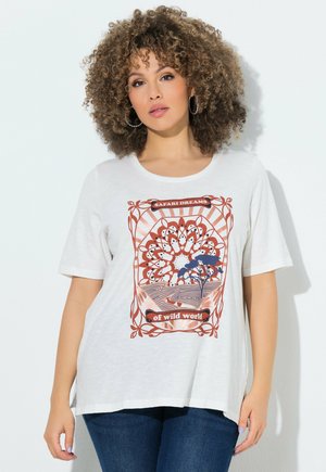 Donna con capelli ricci che indossa una maglietta bianca con la scritta "Safari Dreams of wild world" e jeans blu, in piedi davanti a uno sfondo bianco.