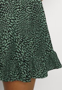 Ourlet de jupe avec motif de feuilles vertes sur tissu noir, présentant un bord froncé au-dessus de la jambe d'une personne.