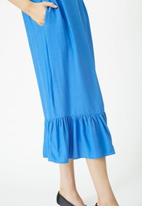 Mėlyna kelių ilgio suknelė su plačiu ruffle hem ir šoninėmis kišenėmis, pagaminta iš minkštos medžiagos su lygiu paviršiumi.