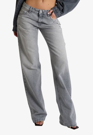 Donna con jeans grigio chiaro a vita alta e gamba larga con leggere abrasioni e tacchi neri con punta quadrata, mano in tasca, addome visibile.