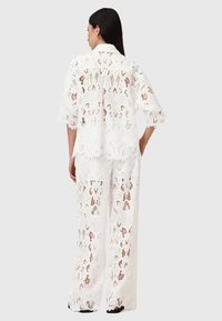 Valkoinen pitsinen kaksiosainen asu, jossa on lyhythihainen paita, kaulus, avoin kudontakukkakuvio ja löysät leveälahkeiset housut.