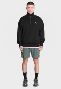 Młody mężczyzna stoi przodem do kamery, ubrany w czarną bluzę na zamek, zielone szorty i trampki, z tatuażami widocznymi na ramionach.