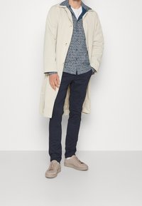 Beige trenčkot přes vzorovanou modrou košili, námořnické kalhoty a světle hnědé tenisky. Viditelné jsou hladké textury a detaily knoflíků.