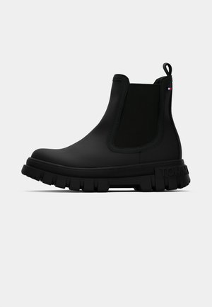 Zwarte rubberen enkellaars met een gestructureerd elastisch paneel, een dikke profielzool en een trekkoord voor gemakkelijke aan- en uittrekken, gebrandmerkt met "TOMMY."