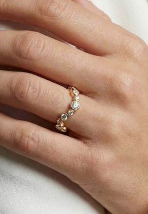 Hånd iført en guldring med syv runde diamanter i forskellige størrelser arrangeret i en buet linje på ringfingeren.