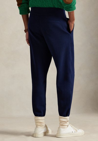 Persoon draagt marineblauwe joggingbroek, witte sneakers met crèmesokken en een groene trui met elleboogstukken, staand met de handen in de zakken.