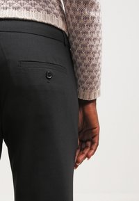 Pantaloni neri con una texture liscia, dotati di una tasca posteriore e dettagli con bottone, abbinati a un maglione lavorato a maglia con motivo in tonalità chiare.