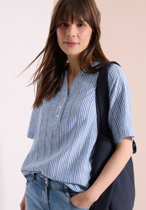 Kobieta o brązowych włosach, ubrana w niebiesko-biały pasiasty t-shirt z krótkim rękawem, niebieskie jeansy, z jedną ręką w kieszeni i granatową torbą na ramieniu.